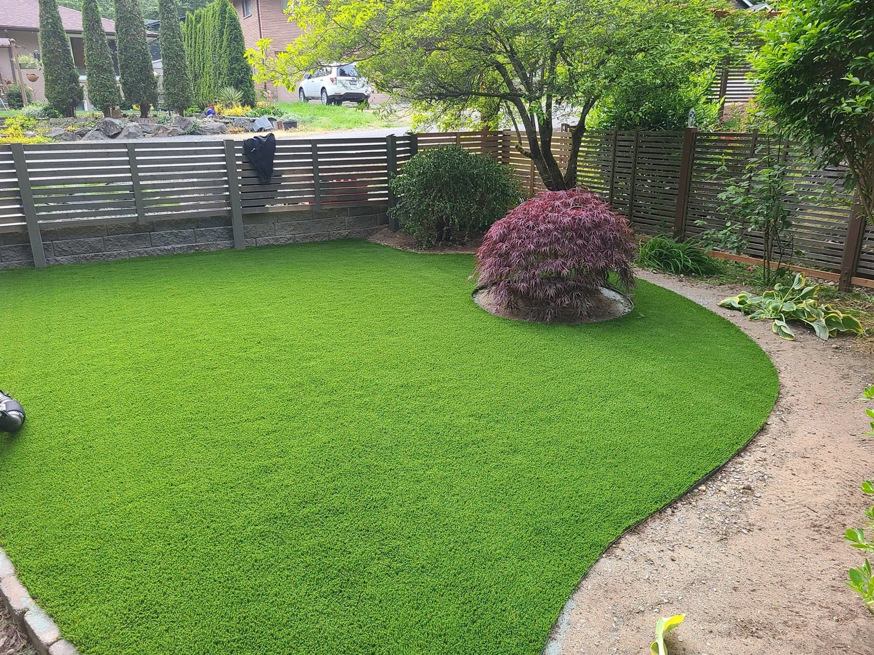 Curved backyard artificial lawn bordered by garden beds, trees, and a privacy fence
