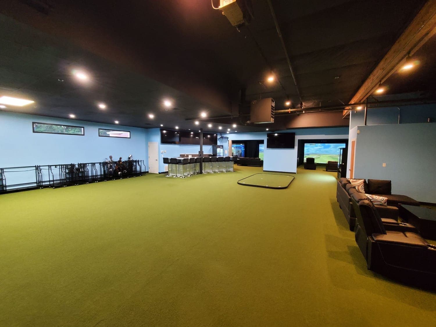 Indoor golf simulator lounge floored with putting turf, club racks, and lounge seating